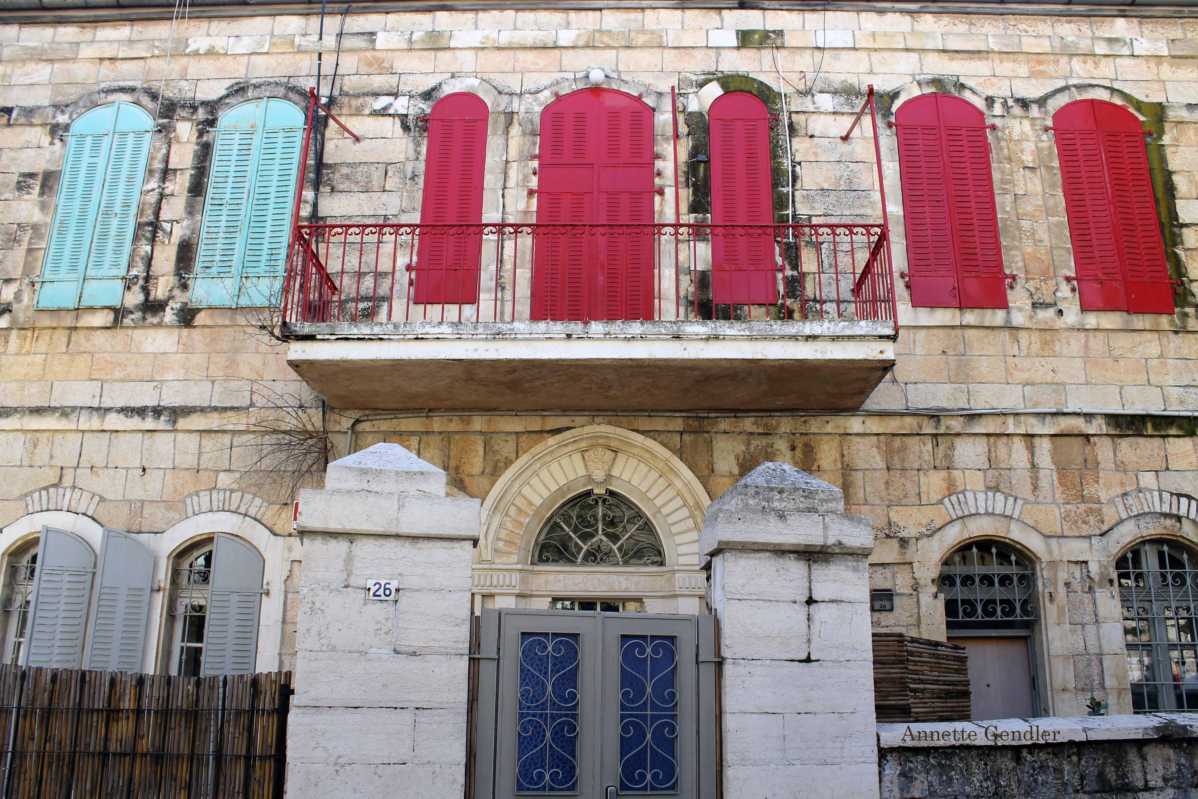 Frontal view of old two-story house in Musrara with red, blue and grey shutters on arched windows