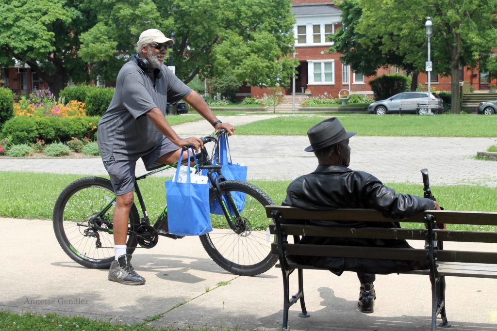Pullman residents, one on a bike, with two shopping bags hanging off its handles, the other sitting on a bench, having a chat in a park