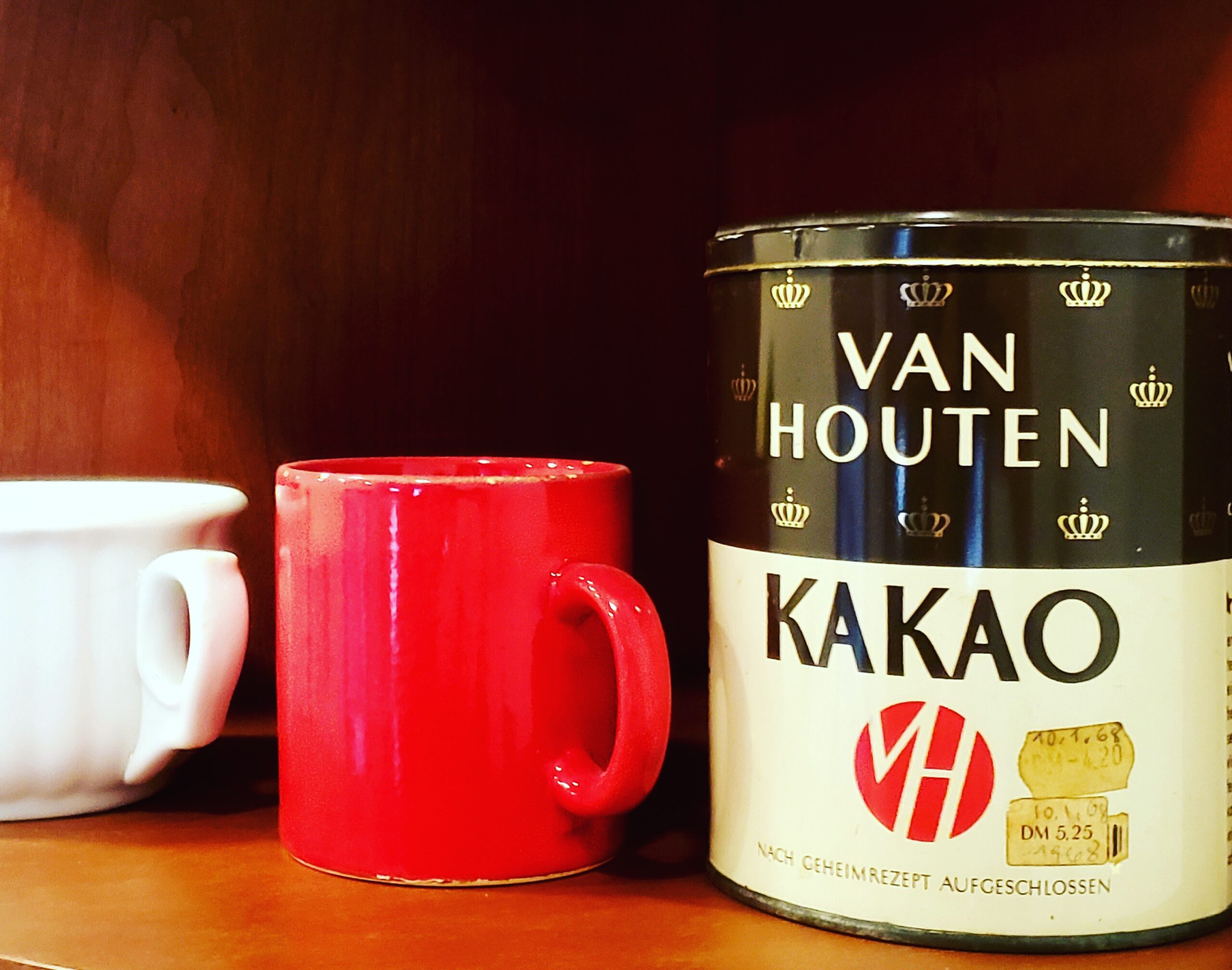 Van Houten cocoa tin on brown shelf with red and white mugs next to it